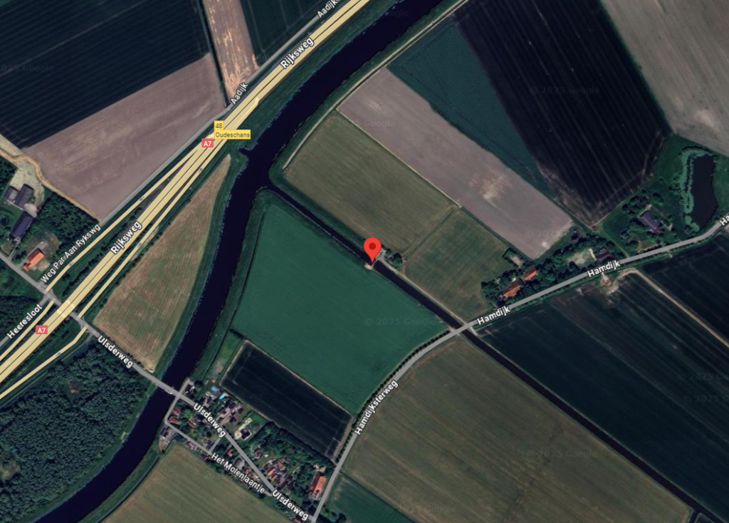 Luchtfoto van het buitengebied nabij Bad Nieuweschans, met centraal op de afbeelding een rood merkteken ter aanduiding van het perceel grond bij poldergemaal Ulsda, gelegen aan de Hamdijk. De exacte locatie bevindt zich ten noorden van het dorp en ten zuiden van de Rijksweg A7, vlak bij afrit 48 (Oudeschans). Het gemarkeerde perceel van circa 24 m² ligt aan een landbouwperceel en direct naast de watergang, in de nabijheid van Hamdijk 53. De afbeelding toont het landelijke karakter van de omgeving met akkers, sloten en verspreide bebouwing.